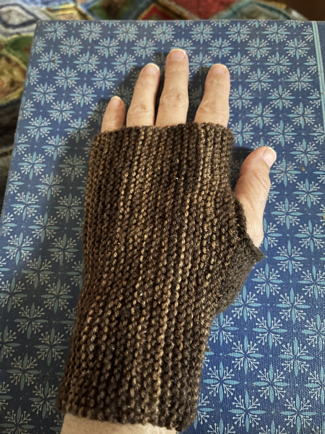 A white person's hand wearing a brown variegated fingerless mitt. It covers the wrist and the main part of the hand leaving the fingers and thumb bare. The tubes covering the hand has vertical ridges that stretch to fit the hand.